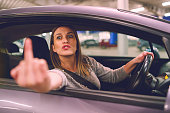 Young woman driver showing middle finger trough the window of the car mad angry furious pissed on the parking in the city at night driving