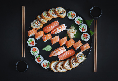 Japanese cuisine. Set  with classic nigiri sushi, fusion hot uramaki, Philadelphia and Unagi rolls from fresh ingredients on round wooden plate at black background. Top view, copy space.
