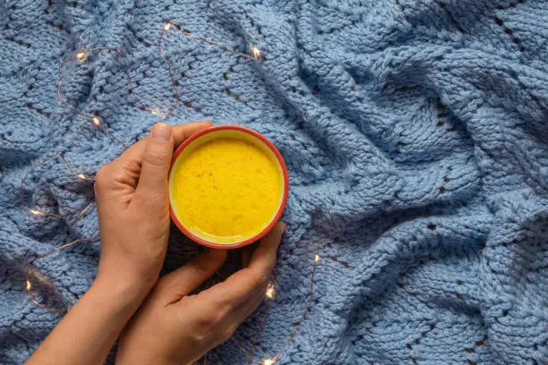 Photo of Woman hands holding a mug of homemade wam turmeric golden milk on blue knitted background with fairy lights.