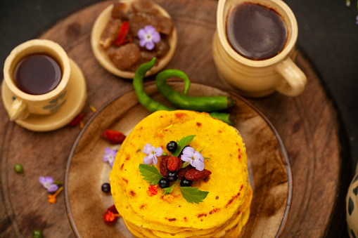 Close-up image of Chilla, Besan Cheela pancakes garnished with flowers, berries, leaf and green chili in plate placed on round wooden try with kettle and cup.