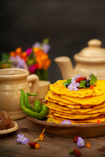 Close-up image of Chilla, Besan Cheela pancakes garnished with flowers, berries, leaf and green chili in plate placed on wooden table with kettle and cup.