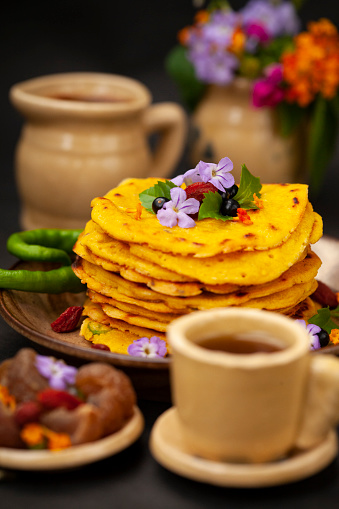 Close-up image of Chilla, Besan Cheela pancakes garnished with flowers, berries, leaf and green chili in plate placed on table with kettle and cup.