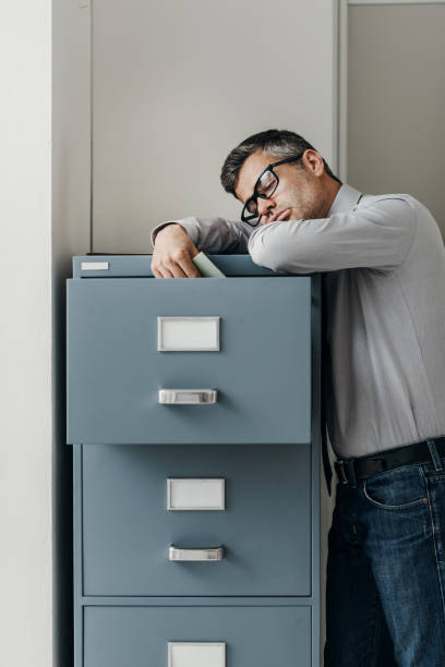 Tired office worker sleeping in the office Tired lazy office worker leaning on a filing cabinet and sleeping, he is falling asleep standing up; stress, unproductivity and sleep disorders concept narcolepsy stock pictures, royalty-free photos & images