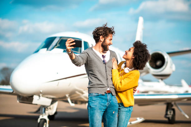 pareja joven rica tomando un selfie en un teléfono móvil frente a un jet privado estacionado en un asfalto del aeropuerto - status symbol audio fotografías e imágenes de stock