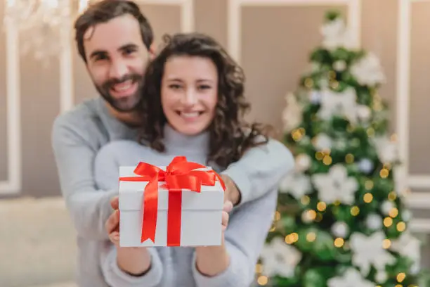 Photo of Defocused man and woman holding a present tied with a red ribbon, looking at the camera smilling.