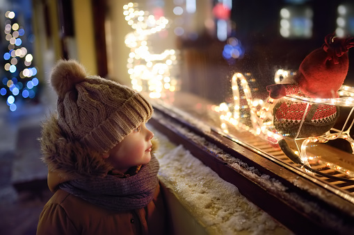 Little boy admires Christmas decorations in showcase of shop on winter evening. Tourist looking on Xmas toys and accessories on traditional Christmas market in Tallinn, Estonia.