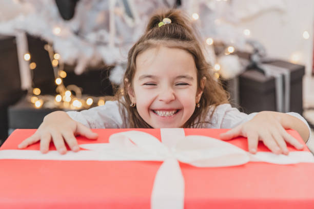 リビングルームの床に座っている間、大きなクリスマスプレゼントを開く超興奮した若い女の子のクローズアップ写真。 - praying joy indoors lifestyles ストックフォトと画像