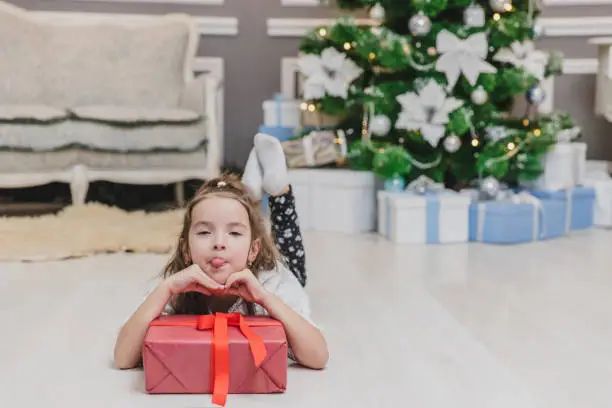 Photo of Closeup of beautiful little angel girl making grimaces, laying on the floor, keepin hands on big present box with ribbon.
