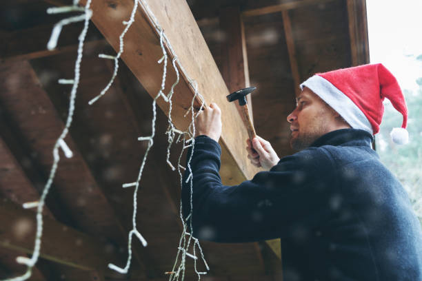 クリスマスの文字列ライトで家の屋外カーポートを飾るサンタ帽子を持つ男 - 吊るす ストックフォトと画像