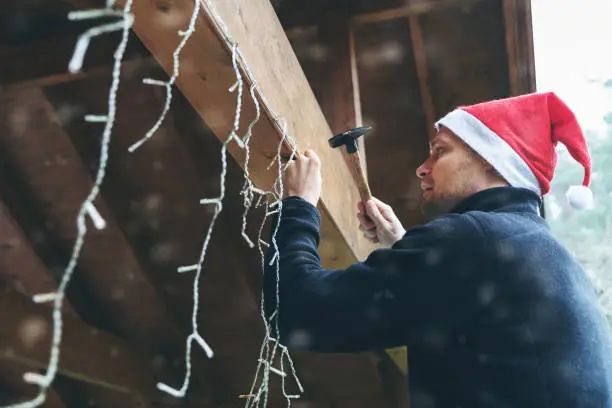 Photo of man with santa hat decorating house outdoor carport with christmas string lights