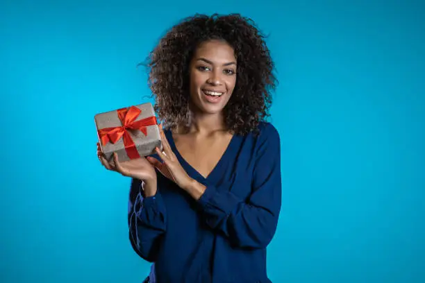 Pretty woman received gift box and interested in what's inside. She is happy and flattered by attention. Girl in Santa hat on blue background. Studio picture