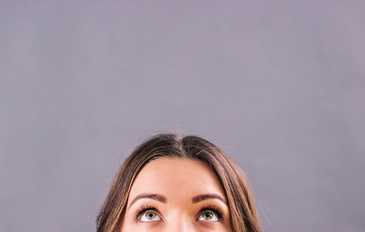 Closeup beautiful young woman looking up on blank copy space isolated on gray background. Attractive funny girl staring above her head. Half face of adorable woman thinking about solution. Empty space