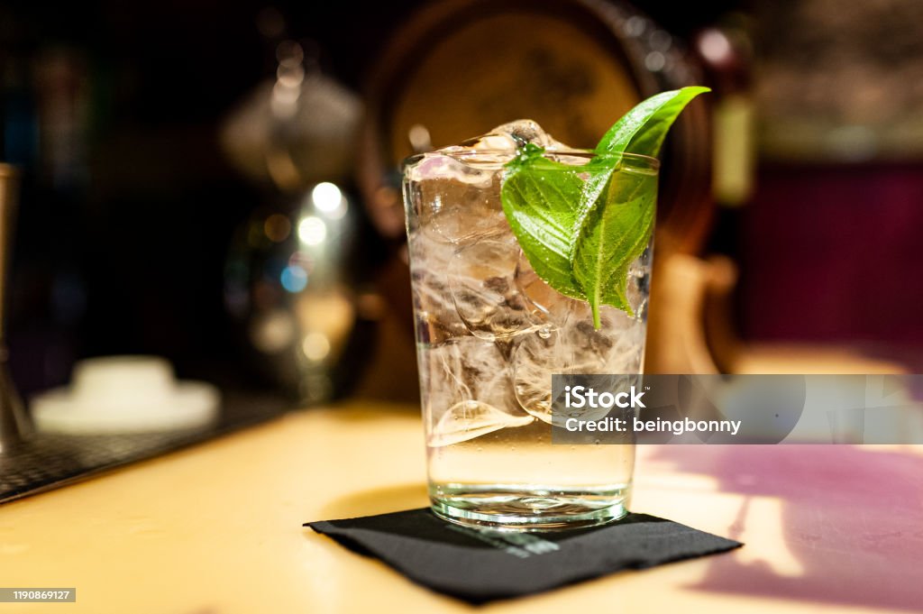 close up of gin tonic glass inside bar on top of bar counter with ice cubes and basil leaves at night Gin Tonic Stock Photo
