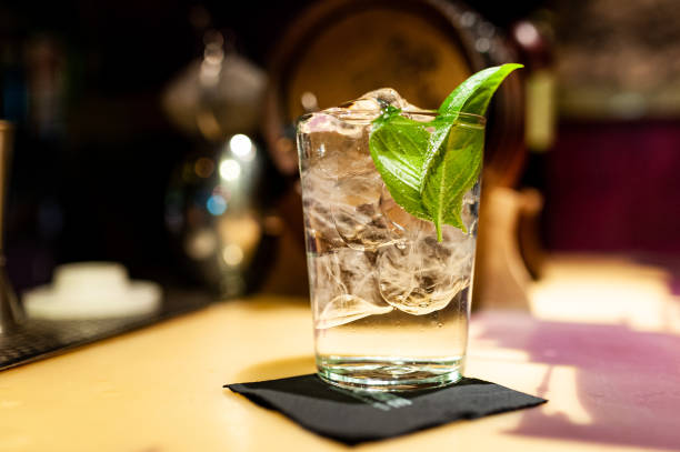 close up of gin tonic glass inside bar on top of bar counter with ice cubes and basil leaves at night stock photo