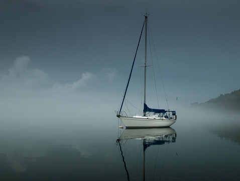 Dramatic single sailboat and its reflection in calm clear water as fog roles in before a storm