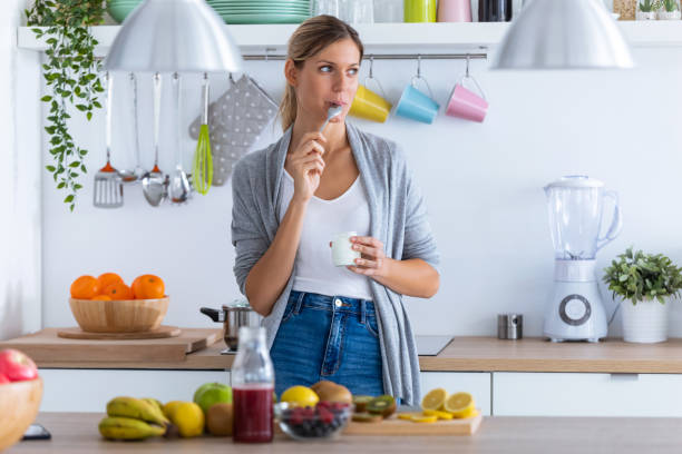 jolie jeune femme mangeant le yaourt tout en restant dans la cuisine à la maison. - routine foods and drinks clothing household equipment photos et images de collection