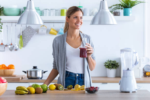 hübsche junge frau, die seitwärts schaut, während sie fruchtsaft in der küche zu hause trinkt. - routine foods and drinks clothing household equipment stock-fotos und bilder