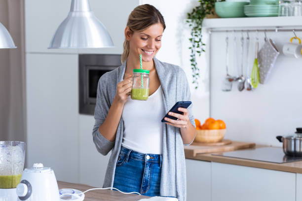 istock Pretty young woman using her mobile phone while drinking detox juice in the kitchen at home. 1190896817