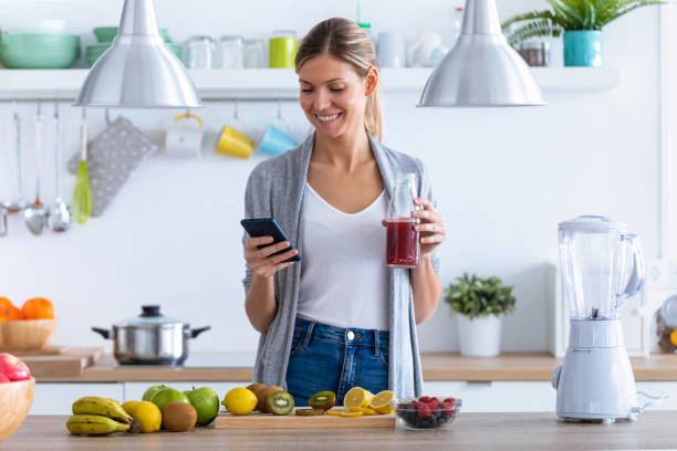 jolie jeune femme utilisant son téléphone portable tout en buvant le jus de fruit dans la cuisine à la maison. - routine foods and drinks clothing household equipment photos et images de collection