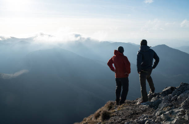 istock Two men standing standing with trekking poles on cliff edge and looking at sunset rays over the clouds. Successful summit concept image. 1190924367