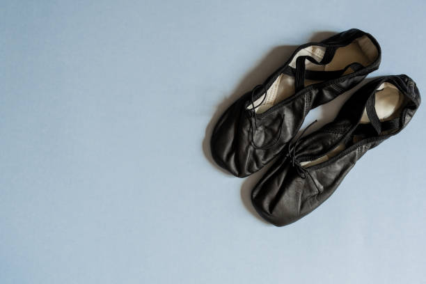 Young adult female ballerina with brunette hair and red flower accessory sits on studio floor. Dressed in black attire and pointe shoes, she exudes elegance. Soft lighting enhances serene atmosphere