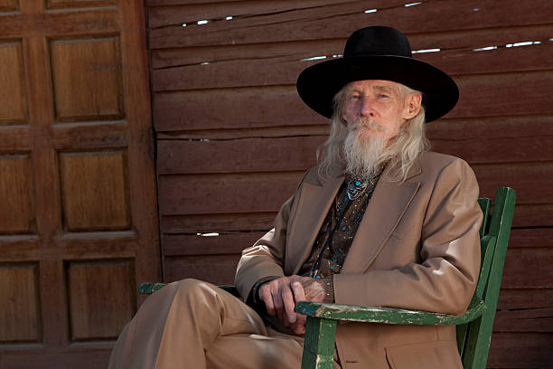 A mexican senior man leaning on a horse box with his arms crossed, thinking and looking away.