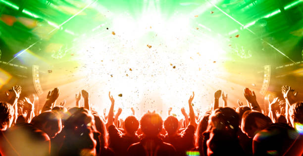 Concert stage, people are visible waving and clapping, silhouettes are visible