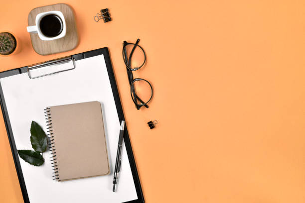 istock Office desk workspace with blank clip board, office supplies, pen, cactus, green leaf, coffee cup on a wooden stand and eye glasses on orange background. Flat lay, top view blog concept 1191008688