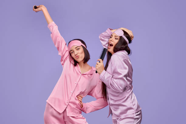 Best friends dancing and singing into comb Excited young women in sleepwear dancing and singing into comb while having fun during sleepover against violet background pyjamas stock pictures, royalty-free photos & images