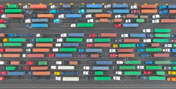 AERIAL, TOP DOWN: Couple of trucks get to drive past the long line waiting in the busy harbor. Flying above a few loaded cargo trucks driving past a queue of others waiting in the Port of Los Angeles.