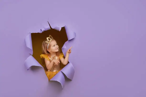 Photo of Cute little child girl in princess costume breaks through a colored purple paper wall. Points with a magic wand to the empty space on the right. Toddler funny emotions face.