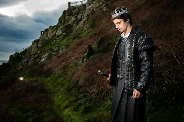 Photo of Handsome king with sword stands in contemplation with hill and parts of keep in the background