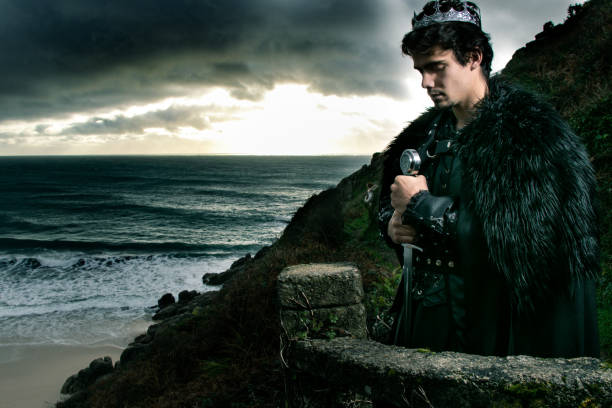 handsome king with goatee stands on stone balcony with sword and crown, praying with ocean in background - feudalism imagens e fotografias de stock