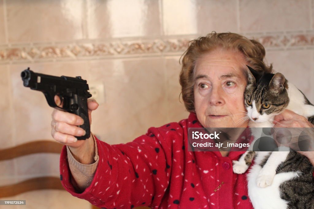 Angry senior woman protecting her cat with a gun Angry senior woman protecting her cat with a gun. Domestic Cat Stock Photo