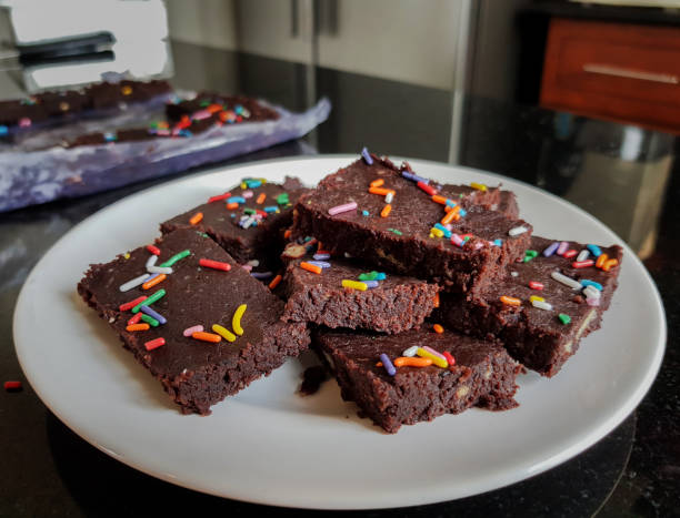 Side angle photo of stacked pieces of Chocolate Biscuit Sprinkle Cakes arranged on a white plate and prepared pieces in the background on a kitchen top stock photo