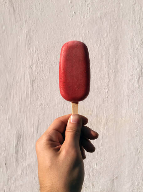 Hand With Red Popsicle Hand holding a red popsicle ice pop on a stick against a solid white wall. popsicle stick stock pictures, royalty-free photos & images