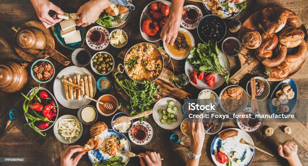 Traditional Turkish breakfast and people taking various food, wide composition Turkish breakfast table. Flat-lay of peoples hands taking pastries, vegetables, greens, olives, cheeses, fried eggs, spices, jams, honey, tea in copper pot and tulip glasses, wide composition Food Stock Photo