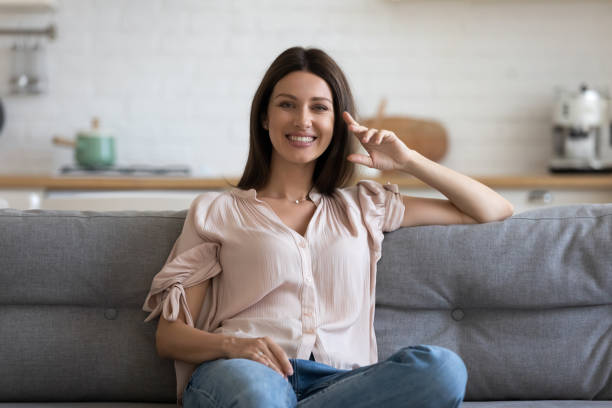 glückliche attraktive junge frau sitzt auf dem sofa und schaut in die kamera - stereotypical housewife women domestic kitchen brown hair stock-fotos und bilder