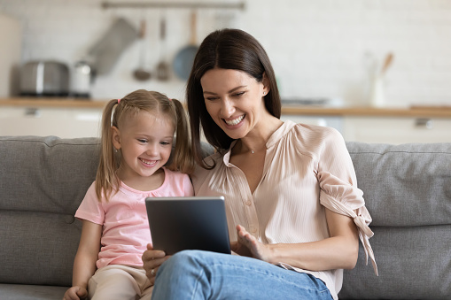 Cheerful young mom with cute little kid daughter using digital tablet computer modern technology app for entertainment education children development doing online video chat relaxing on couch at home