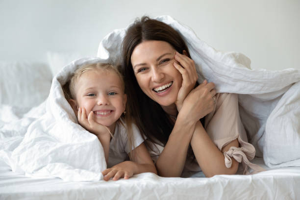 familia feliz mirando la cámara cubierta con edredón blanco cálido - sheet sleeping women bed fotografías e imágenes de stock