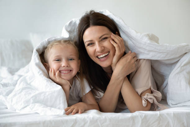 famiglia felice che guarda la macchina fotografica coperta di piumino bianco caldo - sheet women bed sleeping foto e immagini stock