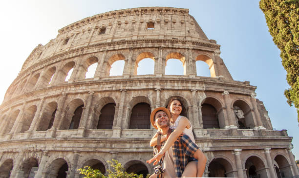 joven pareja feliz divirtiéndose en el coliseo de roma. piggyback posando para fotos. - couple vacations travel destinations europe fotografías e imágenes de stock