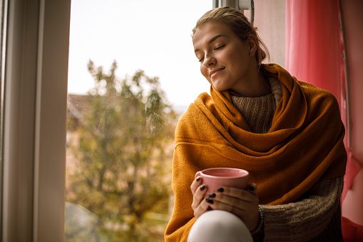 Copy space shot of young woman wrapped in a cozy yellow scarf, sitting on the window cell, with her eyes closed and enjoying a warm cup of tea on a cold autumn day.