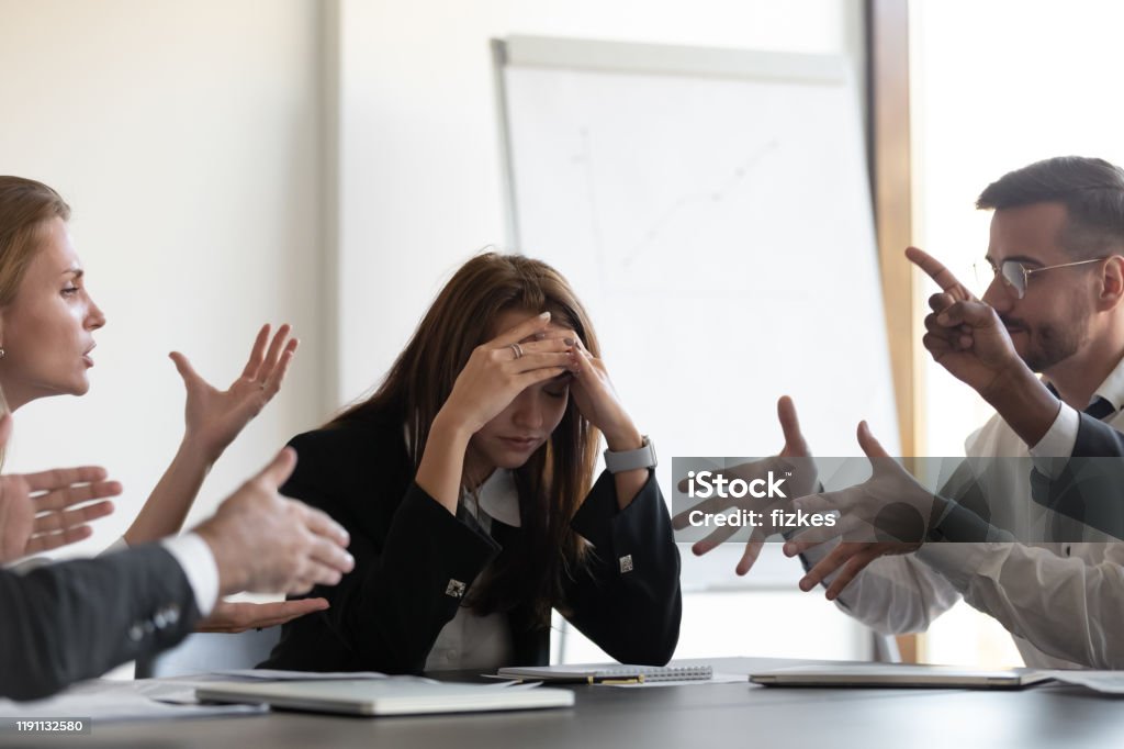 Frustrated millennial female worker felling tired of working quarreling. Frustrated millennial female worker sitting at table with colleagues, felling tired of working quarreling at business meeting. Upset stressed young businesswoman suffering from head ache at office. Conflict Stock Photo