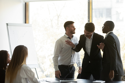 Millennial businessman setting apart two aggressive multiracial coworkers, starting fighting at office. Mixed race young colleagues quarreling shouting at each other, misbehaving during meeting.