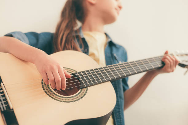 Low angle of anonymous child in casual outfit playing guitar during lesson in music school against beige wall