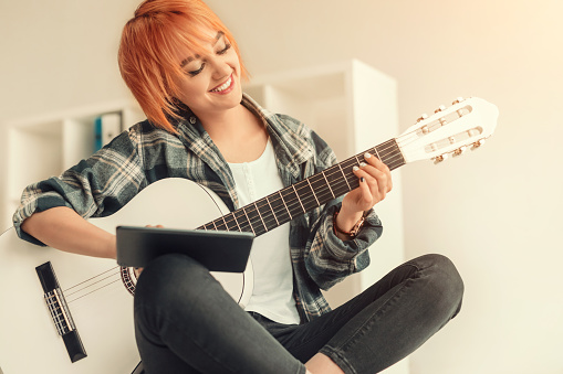 Delighted lady smiling and using tablet while sitting crossed legged and learning to play acoustic guitar in cozy home