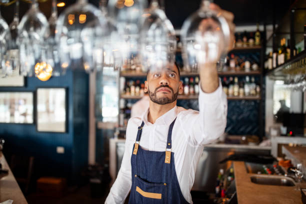 Bartender working at the cafe Mixed race barman working at the cafe. Bartender taking a wineglasses from the overhead rack at bar counter. bar catering stock pictures, royalty-free photos & images