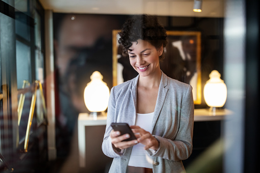 Young businesswoman standing inside a hotel lobby using mobile phone. Female standing at hotel hallway booking online taxi using smart phone app.