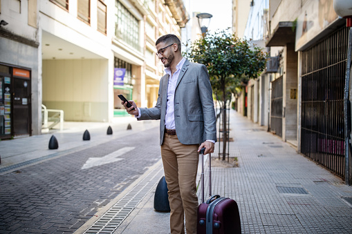 Male executive standing outside a hotel looking at his mobile phone. Businessman with luggage waiting for the taxi to arrive.